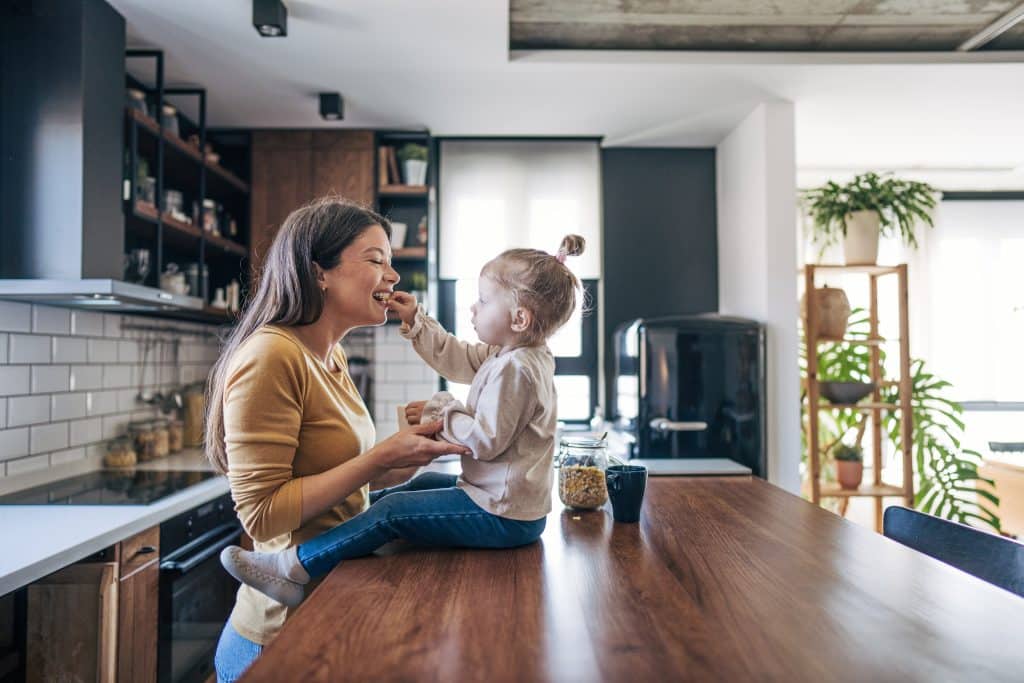 toddler feeding mom while sitting on wood top kitchen island in renovated but homey kitchen